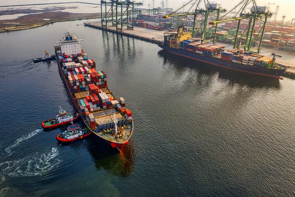 Photo of a container ship being pushed by tug boats toward a maritime port. 